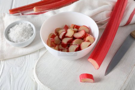 Sliced rhubarb in bowl and on board, knife and bowl of salt on white wooden background, close up