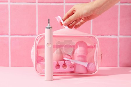 Téléchargez les photos : Brosse à dents électrique, sac cosmétique et main féminine sur fond rose - en image libre de droit