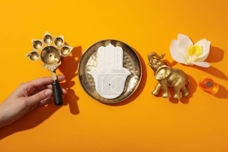 Photo for Copper utensils in hand, elephant figurine, Hamsa and flower on orange background, top view - Royalty Free Image