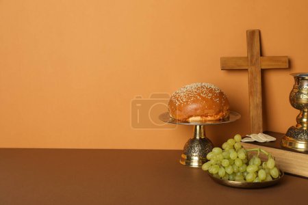 Wooden cross and cup on book, bread and grapes on orange background, space for text