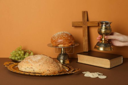 Wooden cross on book, cup in hand, bread and grapes on orange background