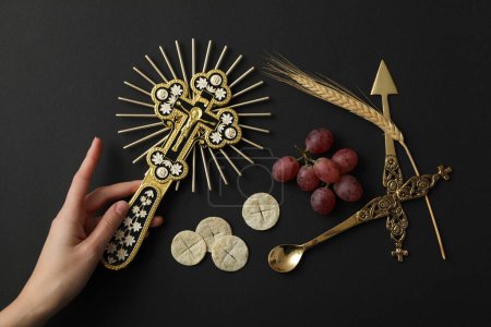 Cross in hand, liturgical bread, grape and cutlery on black background, top view