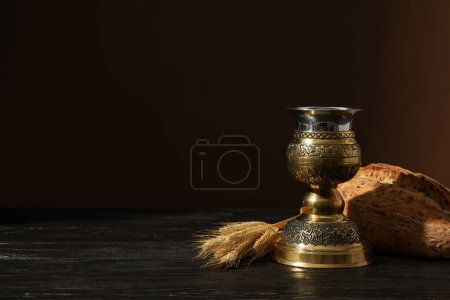 Wooden cross, bread, spikelets and cup on brown background, space for text