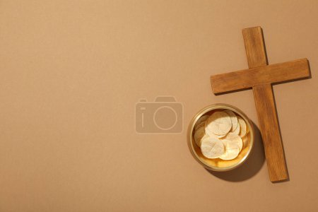 Wooden cross and liturgical bread on brown background, space for text