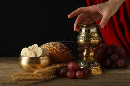 Foto de Copa en mano, pan, espiguillas, uvas sobre mesa de madera sobre fondo negro - Imagen libre de derechos