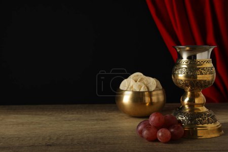 Foto de Pan litúrgico, uvas y copa sobre mesa de madera sobre fondo negro, espacio para texto - Imagen libre de derechos