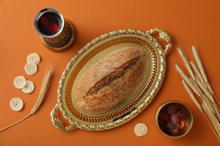 Bread on golden tray, spikelets, grapes and cup of wine on orange background, top view