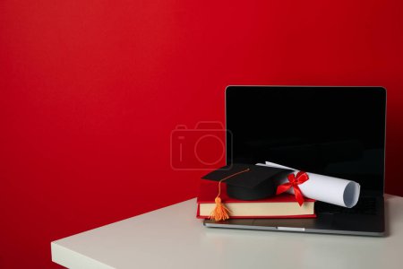 Chapeau diplômé avec diplôme, livre et ordinateur portable sur fond rouge.