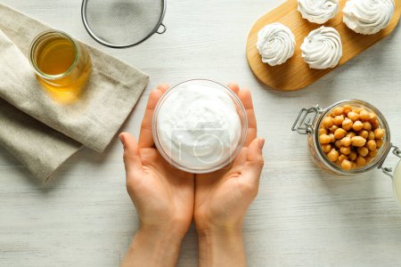 Bowl with cream in hands, jar with chickpeas and aquafaba, marshmallows on board on white wooden background, top view