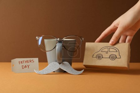 Eine Tasse mit Schnurrbart, ein Geschenk mit Auto und Brille auf orangefarbenem Hintergrund.