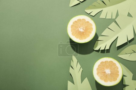 Two half of pomelo fruit, paper tropical leaves on green background, space for text