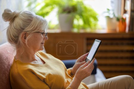 Téléchargez les photos : Femme âgée utilisant e-reader et la lecture d'un e-book à la maison - en image libre de droit