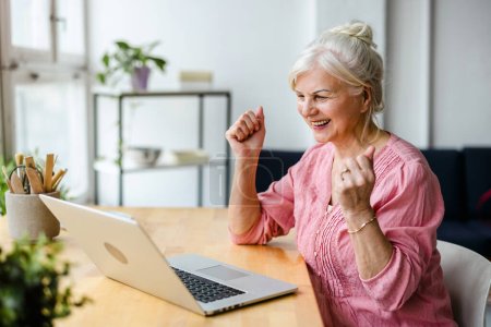 Téléchargez les photos : Femme âgée gaie utilisant un ordinateur portable dans le salon à la maison - en image libre de droit