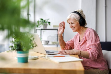 Photo for Happy senior woman in headphones using laptop and waving at camera during video chat - Royalty Free Image