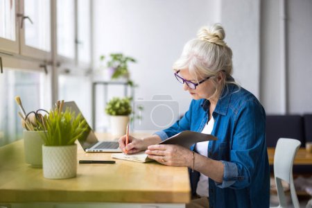 Photo for Smiling mature businesswoman writing in notebook while sitting at table in office - Royalty Free Image