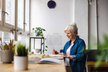 Photo for Smiling mature businesswoman writing in notebook while sitting at table in office - Royalty Free Image