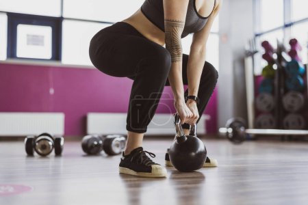 Photo for Young woman lifting Kettle bell while crouching in gym - Royalty Free Image