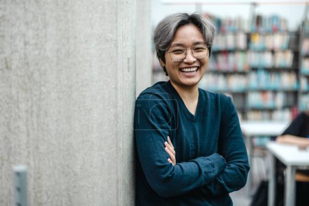 Téléchargez les photos : Portrait d'étudiant asiatique souriant debout avec les bras croisés dans la bibliothèque du collège - en image libre de droit