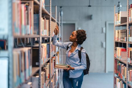 Photo for Black female student picking book from bookshelf in library - Royalty Free Image