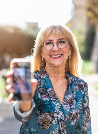 Téléchargez les photos : Portrait de femme âgée souriante avec téléphone portable dans la rue - en image libre de droit