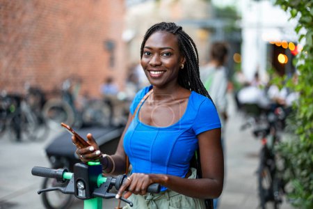 Téléchargez les photos : Portrait de jeune femme heureuse utilisant son smartphone tout en se tenant debout avec scooter électrique dans la ville - en image libre de droit