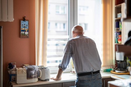Photo for Senior man standing at the kitchen counter in his house and looking out the window - Royalty Free Image