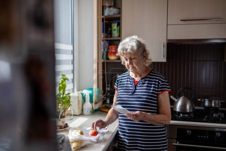 Téléchargez les photos : Femme âgée passant par ses reçus à la maison après avoir acheté des produits d'épicerie - en image libre de droit