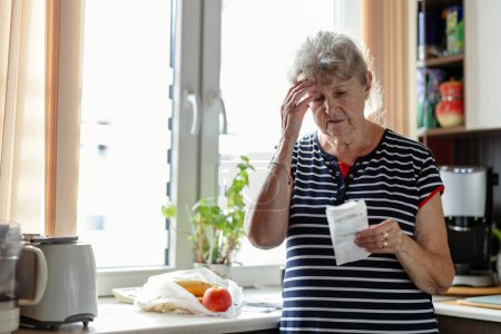Téléchargez les photos : Femme âgée passant par ses reçus à la maison après avoir acheté des produits d'épicerie - en image libre de droit