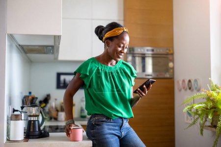 Téléchargez les photos : Femme souriante utilisant un smartphone dans la cuisine à la maison - en image libre de droit