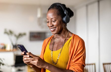 Téléchargez les photos : Femme écoutant de la musique avec des écouteurs connectés à son smartphone dans le salon à la maison - en image libre de droit