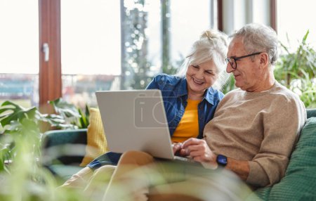 Téléchargez les photos : Couple aîné utilisant un ordinateur portable assis sur le canapé dans le salon à la maison - en image libre de droit