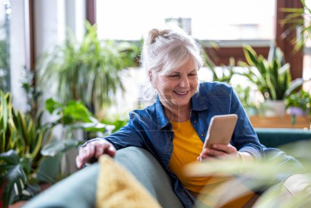 Foto de Mujer mayor sonriente usando un teléfono inteligente mientras está sentada en el sofá en casa - Imagen libre de derechos