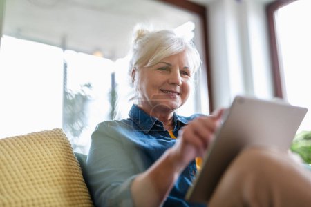 Téléchargez les photos : Femme mûre utilisant une tablette numérique tout en étant assis sur le canapé à la maison - en image libre de droit