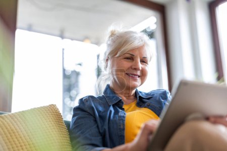 Téléchargez les photos : Femme mûre utilisant une tablette numérique tout en étant assis sur le canapé à la maison - en image libre de droit