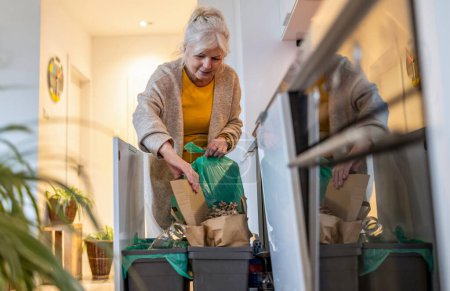 Téléchargez les photos : Femme âgée triant les ordures dans des bacs de recyclage à la maison - en image libre de droit
