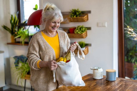 Foto de Mujer mayor sosteniendo bolsa reutilizable con comestibles en la cocina - Imagen libre de derechos