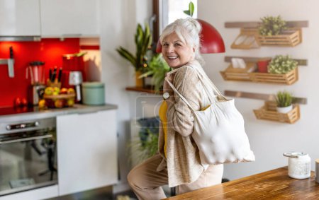 Foto de Mujer mayor sosteniendo bolsa reutilizable con comestibles en la cocina - Imagen libre de derechos
