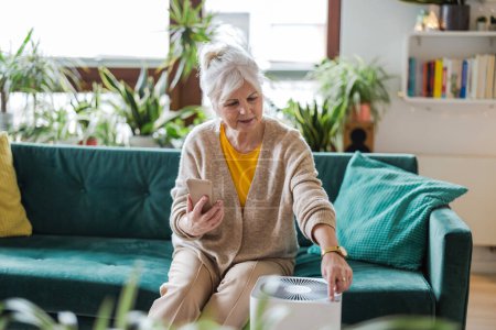 Téléchargez les photos : Femme âgée utilisant un smartphone pour installer un purificateur d'air à la maison - en image libre de droit