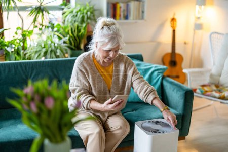 Téléchargez les photos : Femme âgée utilisant un smartphone pour installer un purificateur d'air à la maison - en image libre de droit