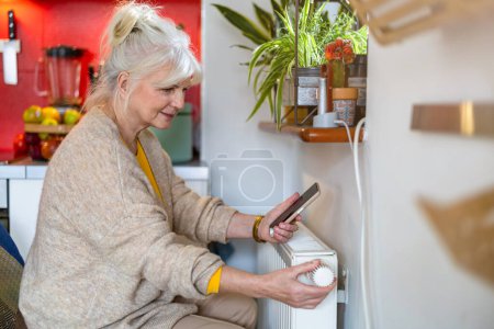 Téléchargez les photos : Femme âgée tenant un téléphone portable tout en ajustant le thermostat sur le radiateur à la maison - en image libre de droit