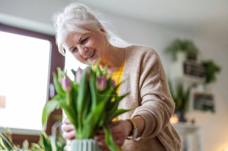 Téléchargez les photos : Heureuse femme âgée avec bouquet de tulipes dans un vase à la maison - en image libre de droit