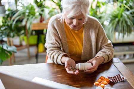 Téléchargez les photos : Femme âgée prenant des pilules d'une bouteille tout en étant assis à la table à la maison - en image libre de droit