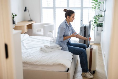 Téléchargez les photos : Jeune femme avec une valise assise sur le lit dans la chambre d'hôtel et utilisant le téléphone - en image libre de droit