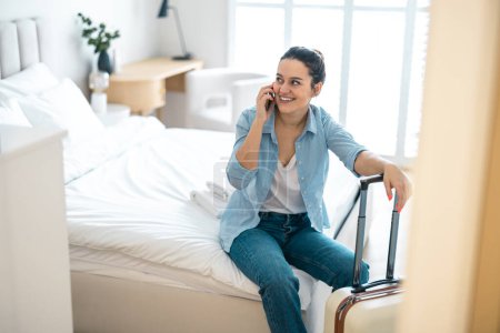Foto de Mujer joven con una maleta sentada en la cama en la habitación del hotel y usando el teléfono - Imagen libre de derechos
