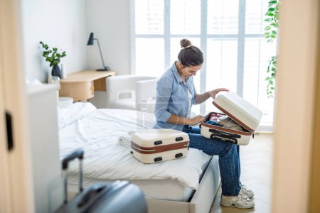 Téléchargez les photos : Jeune femme emballant une valise dans la chambre, se préparant pour le voyage - en image libre de droit