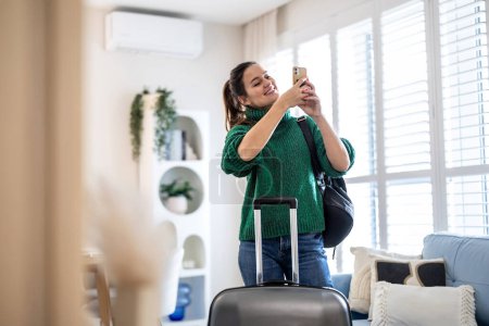 Téléchargez les photos : Jeune femme utilisant un téléphone portable debout avec sa valise dans la chambre d'hôtel - en image libre de droit