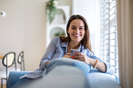 Téléchargez les photos : Femme souriante utilisant un téléphone portable sur le canapé dans le salon à la maison - en image libre de droit