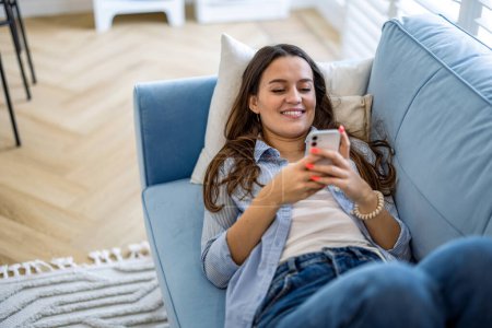 Foto de Mujer sonriente usando teléfono móvil en el sofá en la sala de estar en casa - Imagen libre de derechos