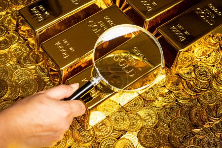 eine Hand mit einer Lupe überprüft den Goldbarren auf einem Haufen Goldmünzen