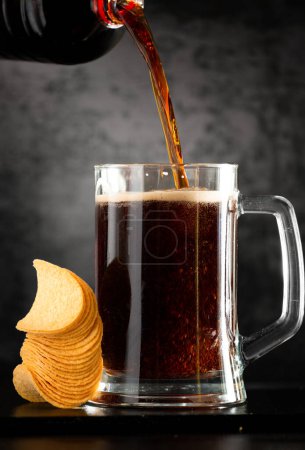 Foto de A glass of dark beer with a handful of chips on a dark background. Beer pouring from a bottle into a glass. Chips in a stack near a mug of beer with foam. - Imagen libre de derechos
